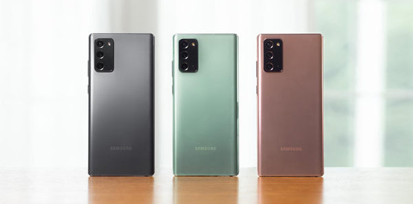 Three Samsung smartphones in black, green, and rose gold on a reflective surface with a blurred background.