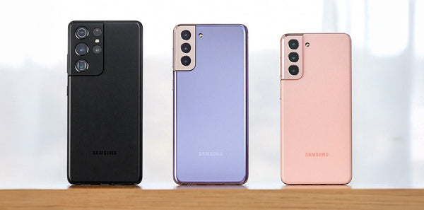 Three Samsung smartphones in black, purple, and pink on a wooden surface with a blurred background.